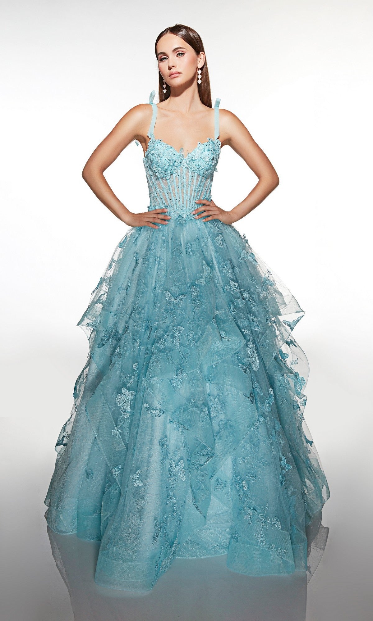 A woman stands gracefully against a plain backdrop wearing the Alyce 62199 long prom dress—a light blue gown with a corset bodice, tulle ballgown skirt, floral appliqués, and delicate spaghetti straps.